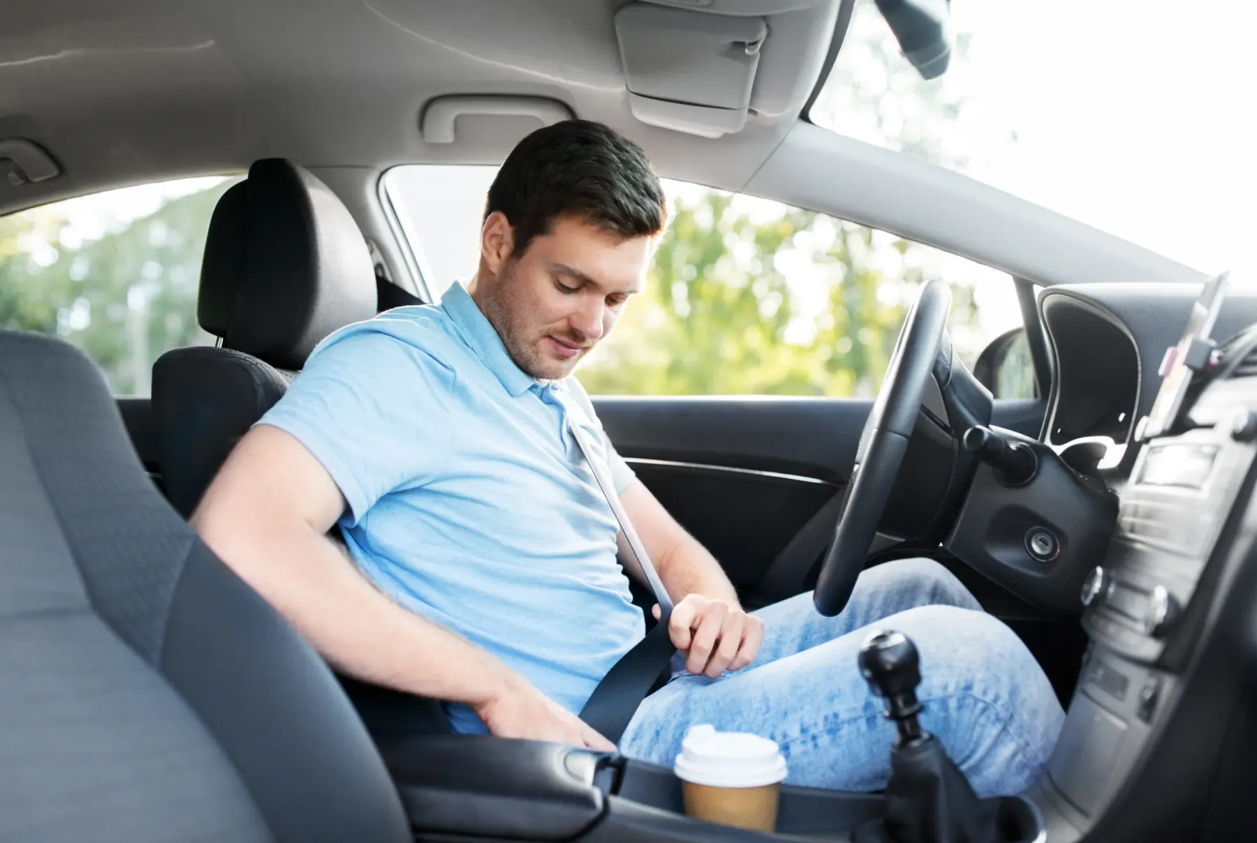 A driver in a passenger car reaching down to fasten the three-point seat belt buckle beside the seat cushion