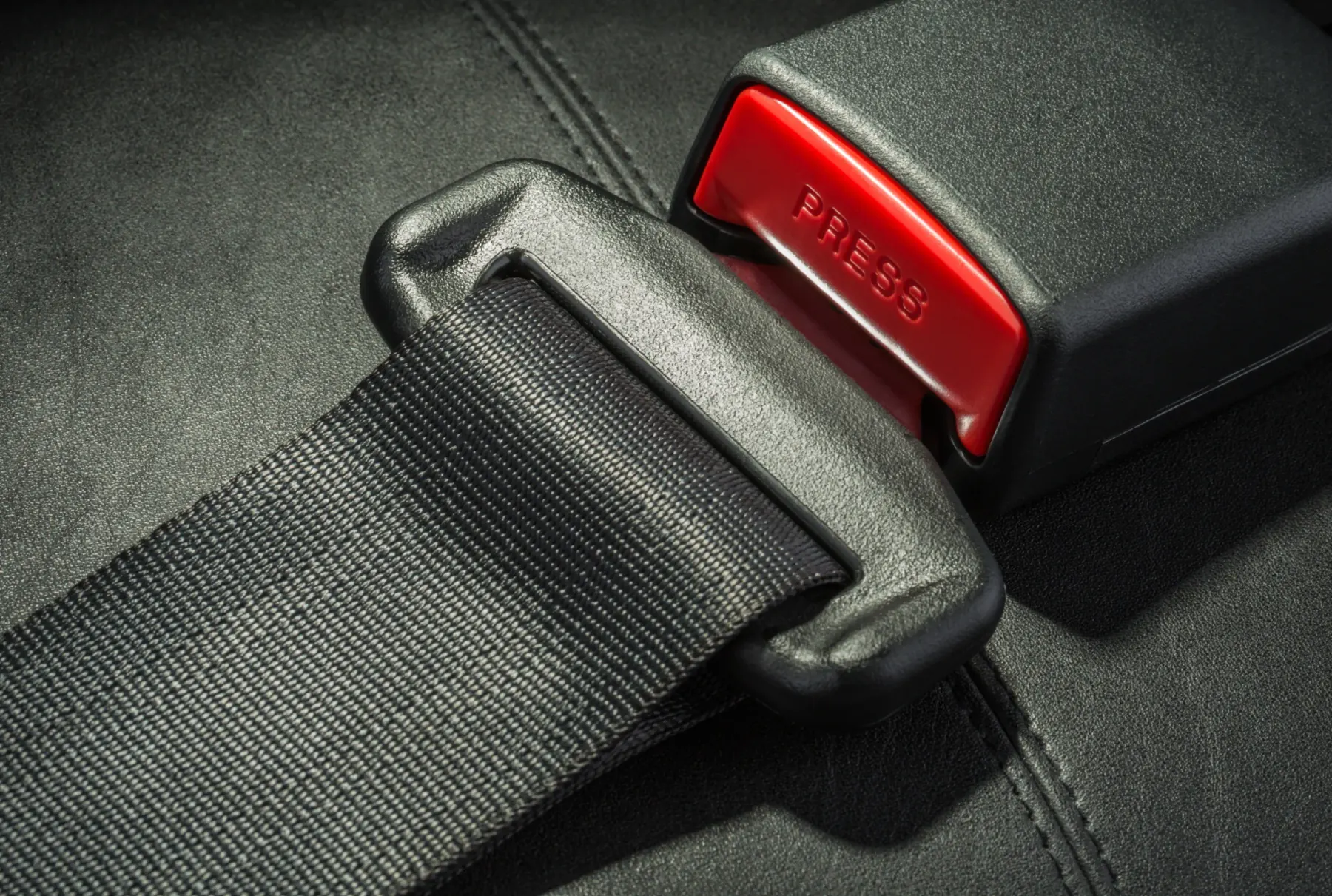 Detailed view of a fastened automotive seat belt with the latch plate seated inside a black buckle housing marked PRESS on a dark leather seat