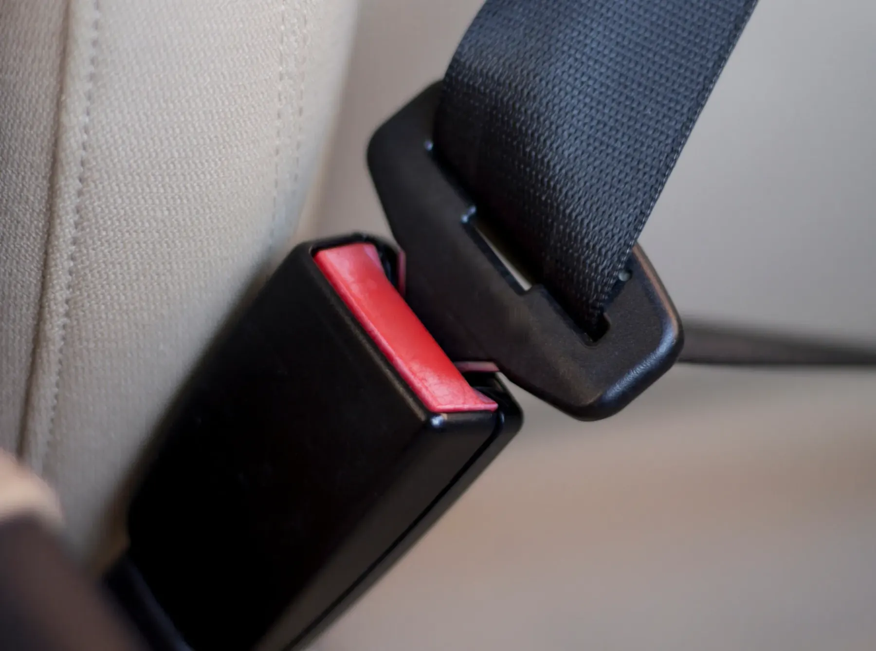 A retractable seat belt latch plate approaching a black buckle receptacle with an exposed red release button, mounted beside a light coloured vehicle seat